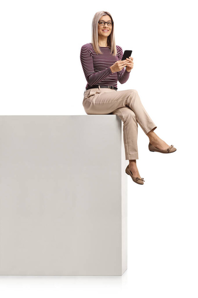 Smiling young woman sitting on a wall and using a mobile phone isolated on white background - Photo, Image
