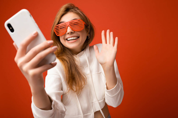 Ελκυστική γοητευτική νεαρή χαμογελαστή ευτυχισμένη γυναίκα κρατώντας και χρησιμοποιώντας το κινητό τηλέφωνο λήψη selfie φορώντας κομψά ρούχα απομονώνονται σε φόντο τοίχο - Φωτογραφία, εικόνα