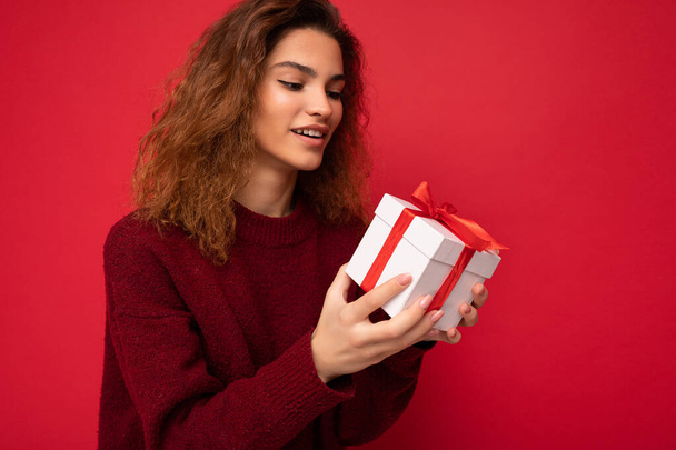 Shot of attractive positive smiling young brunette curly woman isolated over red background wall wearing red sweater holding gift box looking at white box with red ribbon and thinking - Fotografie, Obrázek