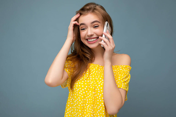 Atrakcyjna pozytywna uśmiechnięta młoda blondynka ubrana w stylowo żółtą letnią sukienkę stojąca samotnie nad niebieskim tłem trzymająca i rozmawiająca przez telefon komórkowy patrząca w bok - Zdjęcie, obraz
