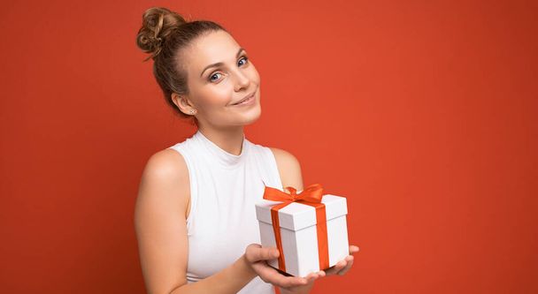 Pretty happy young blonde female person isolated over red background wall wearing white top holding gift box and looking at camera - Foto, Imagem