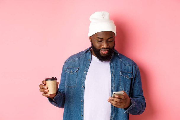 Εικόνα από κομψό μαύρο hipster άνθρωπος πίνοντας καφέ takeaway, ανάγνωση μήνυμα στο τηλέφωνο, στέκεται πάνω από ροζ φόντο - Φωτογραφία, εικόνα