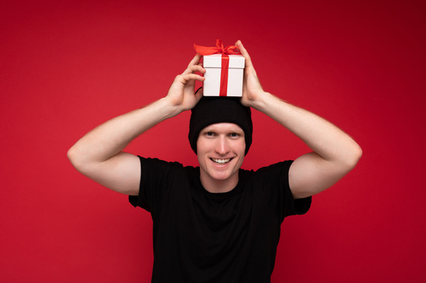 Photo d'un bel homme adulte souriant et positif isolé sur un mur de fond rouge portant un chapeau noir et un t-shirt noir tenant une boîte-cadeau blanche avec un ruban rouge et regardant la caméra et s'amusant - Photo, image
