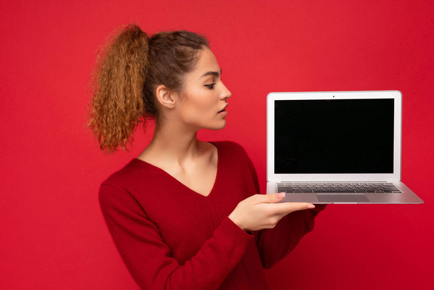 Beautiful charming fascinating young curly dark blond woman wearing red sweater standing isolated over red wall background holding computer laptop with empty copy space mock up pointing to the side - Foto, Imagem