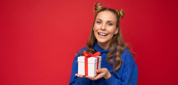 Beautiful happy young blonde woman isolated over colourful background wall wearing stylish casual clothes holding gift box and looking at camera - Photo, image