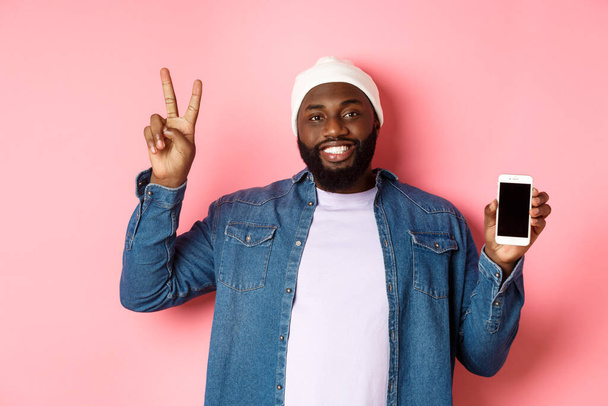 Compras en línea y concepto de tecnología. Chico negro feliz en gorro y camisa de mezclilla mostrando pantalla móvil y signo de paz, de pie sobre fondo rosa - Foto, imagen