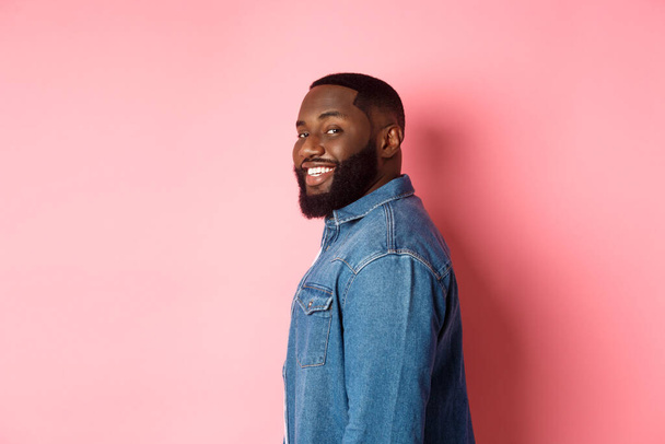 Όμορφος αφροαμερικάνος με γενειάδα, γυρνά το πρόσωπο στην κάμερα και χαμογελά με αυτοπεποίθηση, στέκεται πάνω από ροζ φόντο - Φωτογραφία, εικόνα