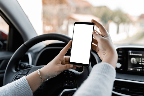 Young driver using smartphon in the car while driving or parking. Mockup of a mobile phone with isolated white screen in the hands of a woman. - Foto, afbeelding