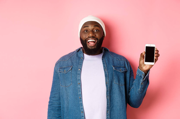 Schöner Hipster in Mütze und Jeanshemd, lächelnd, zeigt Handybildschirm mit glücklichem Gesicht, empfehle App, steht über rosa Hintergrund - Foto, Bild
