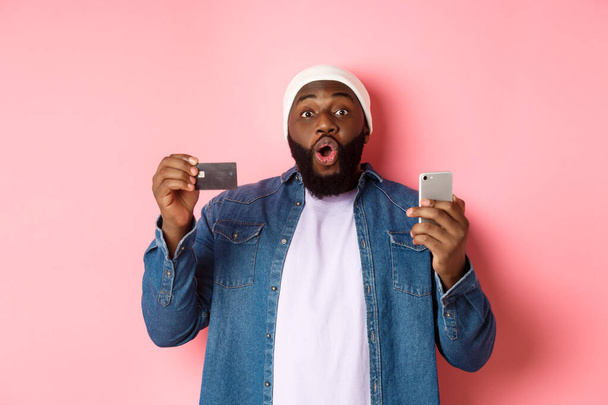 Achats en ligne. Homme noir excité montrant la carte de crédit, en utilisant un smartphone, regardant la caméra étonnée, debout sur fond rose - Photo, image