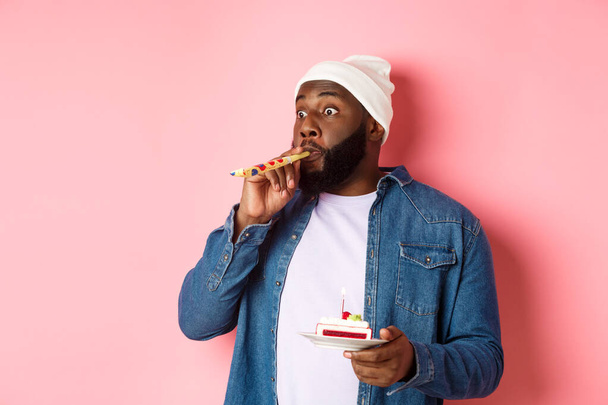 Happy Black hipster celebrating birthday, blowing party whistle, holding bday cake with candle, standing over pink background - Photo, Image