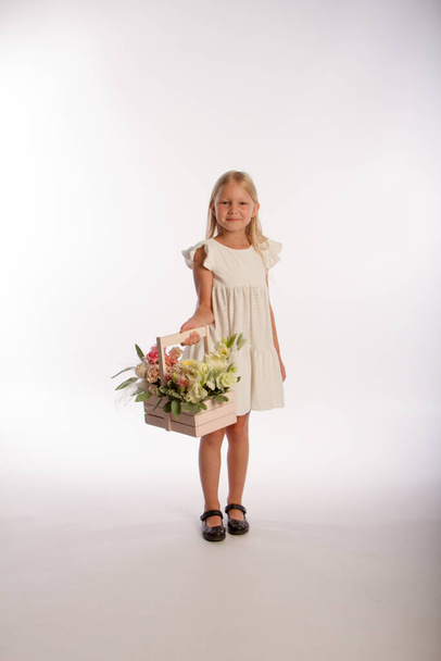 Studio portrait of cute blonde girl in white dress with wooden basket of flowers, white background, selective focus - Foto, Imagen