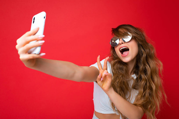 Close-up foto van mooie jonge vrouw met mobiele telefoon het nemen van selfie foto met behulp van smartphone camera dragen zonnebril alledaagse stijlvolle outfit geïsoleerd over kleurrijke muur achtergrond op zoek naar - Foto, afbeelding