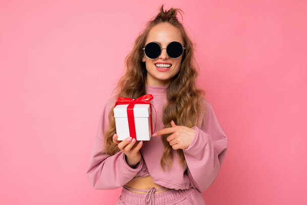 привлекательная счастливая улыбающаяся молодая блондинка кудрявая женщина изолирована на розовом фоне стены в розовой спортивной одежде и солнцезащитных очках держа подарочную коробку глядя на камеру - Фото, изображение