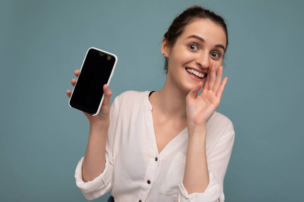 Beautiful positive young blonde curly woman wearing casual white shirt and isolated over blue background wall holding phone and showing smartphone with empty dcreen for mockup looking at camera and - 写真・画像