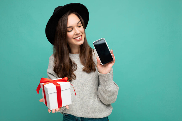 Photo of beautiful happy joyful young brunet woman isolated on blue background wall wearing black hat and grey sweater holding gift box showing mobile phone screen display for mockup and looking at - Photo, Image