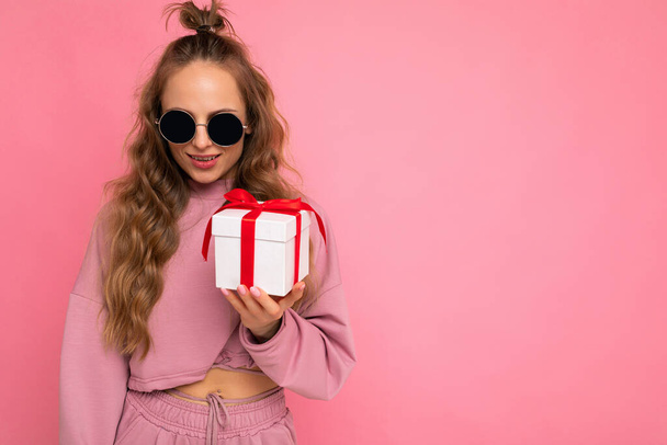 Atractiva mujer rizada rubia joven positiva aislada sobre la pared de fondo rosa que usa ropa deportiva rosa y gafas de sol que sostienen la caja de regalo mirando a la cámara - Foto, Imagen