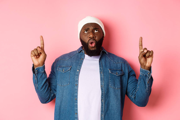 Disappointed african american guy complaining, looking and pointing fingers up at bad promo, standing displeased against pink background - Photo, Image