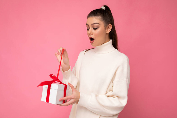 Photo shot of pretty positive surprised young brunet woman isolated over colourful background wall wearing trendy outfit look holding gift box and looking at present box with red ribbon - Foto, immagini