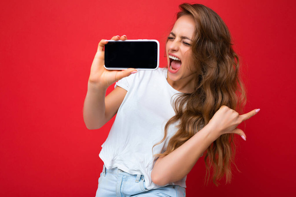 насолоджуючись красивою усміхненою молодою блондинкою, добре виглядає в білій футболці, що стоїть ізольовано на червоному тлі з копією простору, що тримає телефон, що показує смартфон в руці з порожнім дисплеєм екрана
 - Фото, зображення