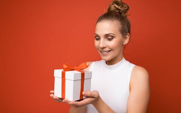 Fénykép lövés gyönyörű pozitív mosolygós fiatal szőke nő elszigetelt felett piros háttér fal visel fehér felső holding ajándék doboz és nézi a jelen - Fotó, kép