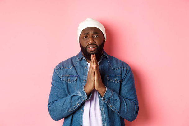 Афро-американський чоловік, який просить допомоги, тримає руки в молитві, дивиться на камеру сумно і молиться, стоїть над рожевим тлом
 - Фото, зображення
