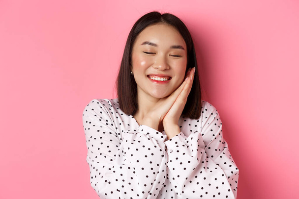Concepto de belleza y estilo de vida. Primer plano de hermosa y soñadora mujer asiática durmiendo sobre sus manos, ojos cerrados y sonriente, soñando despierto sobre fondo rosa - Foto, imagen