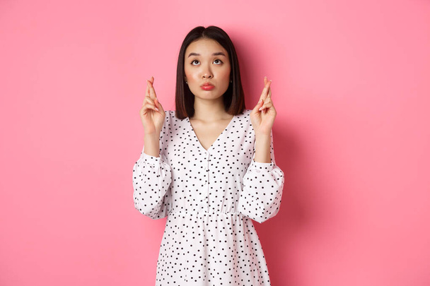 Cute asian woman making a wish, smiling happy and cross fingers for good luck, waiting over pink background - Photo, Image