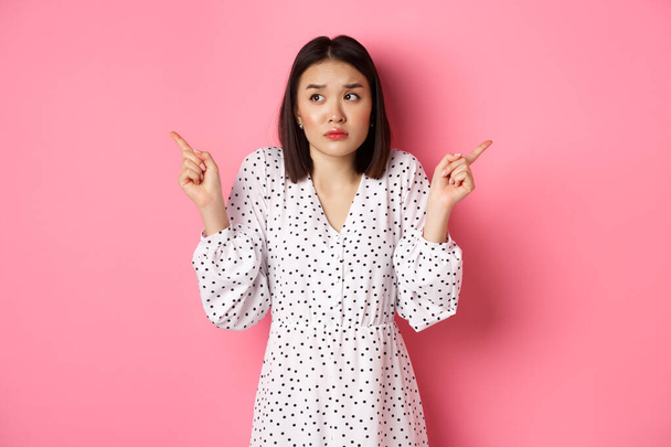 Confused asian woman hesitating, shrugging and pointing hands two sides, staring indecisive and need help with choice, standing over pink background - Photo, Image