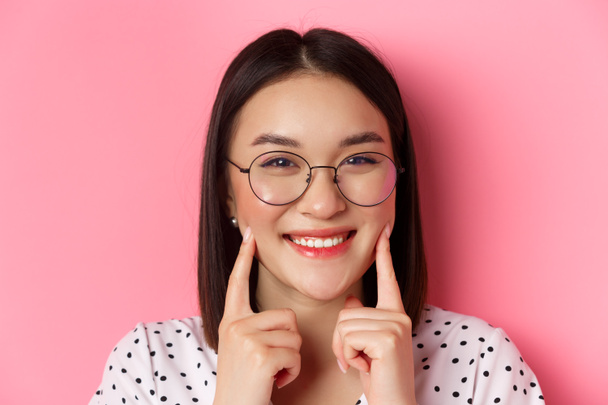 Koncepcja piękna. Zdjęcie głowy uroczej azjatyckiej dziewczyny w modnych okularach uśmiechniętej, szturchającej policzki i pokazującej słodkie dołeczki, stojącej na różowym tle - Zdjęcie, obraz