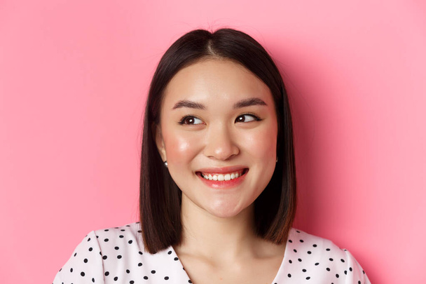 Concept de beauté et style de vie. Headshot de jolie asiatique femme rêveuse regardant gauche à l'espace de copie, souriant heureux, debout sur fond rose - Photo, image