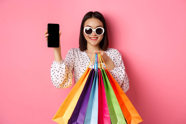 Attractive asian woman showing smartphone app and shopping bags, buying online via application, standing over pink background - Foto, afbeelding