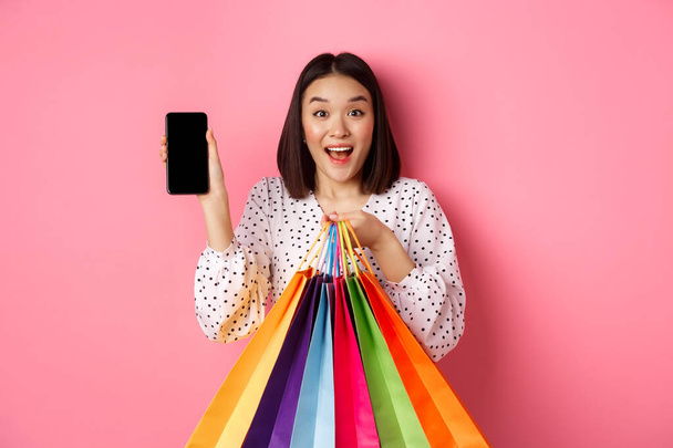 Atractiva mujer asiática mostrando aplicación de teléfono inteligente y bolsas de compras, compra en línea a través de la aplicación, de pie sobre fondo rosa - Foto, Imagen