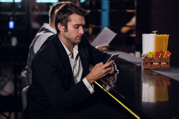 ナイトクラブのバーの後ろのスマートフォンでネットサーフィンをしている美しい若い男 - 写真・画像