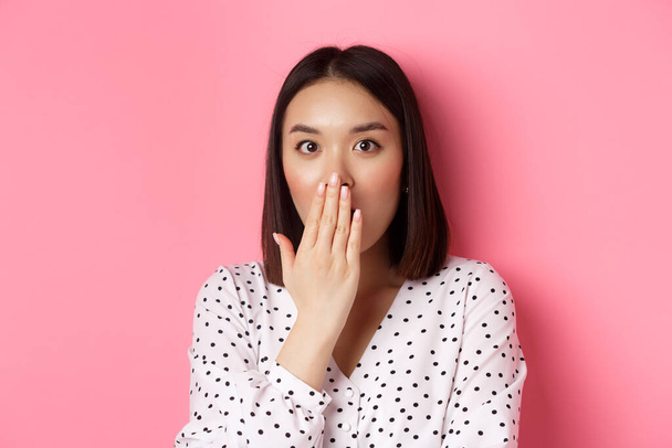 Image d'une fille asiatique choquée bavardant, haletant et couvrant la bouche, regardant la caméra avec une incrédulité totale, debout sur fond rose - Photo, image