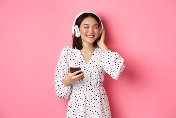 Romantikus ázsiai lány zenét hallgat fejhallgató, mosolygós csukott szemmel, kezében mobiltelefon, álló felett rózsaszín háttér - Fotó, kép