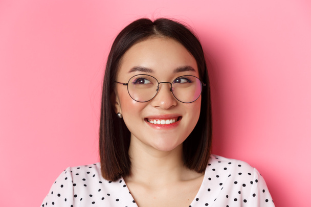 Krása a životní styl. Detailní záběr roztomilé asijské modelky nosí módní brýle, usmívá se a dívá se vlevo na kopírovací prostor, stojí na růžovém pozadí - Fotografie, Obrázek