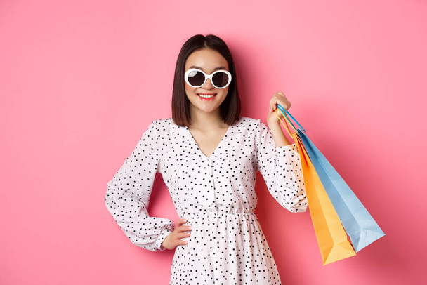 Atractiva mujer asiática sonriente sosteniendo bolsas de compras, usando gafas de sol y vestido lindo, de pie sobre fondo rosa - Foto, imagen