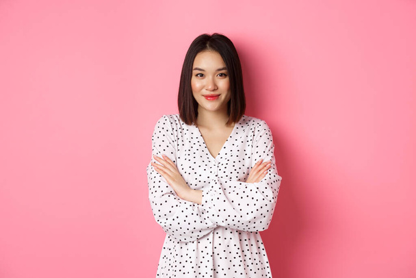 Schönes asiatisches Model im Kleid, verschränkte Arme auf der Brust und lächelnd in die Kamera, vor rosa Hintergrund stehend - Foto, Bild