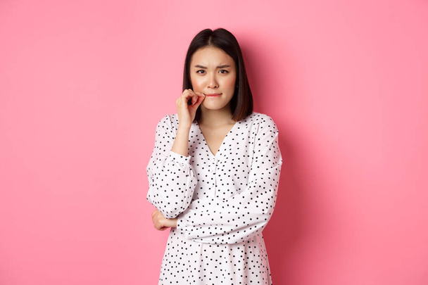 Brunette asian woman in dress looking displeased, frowning and zipping mouth, seal lips with promise, standing over pink background - Фото, зображення