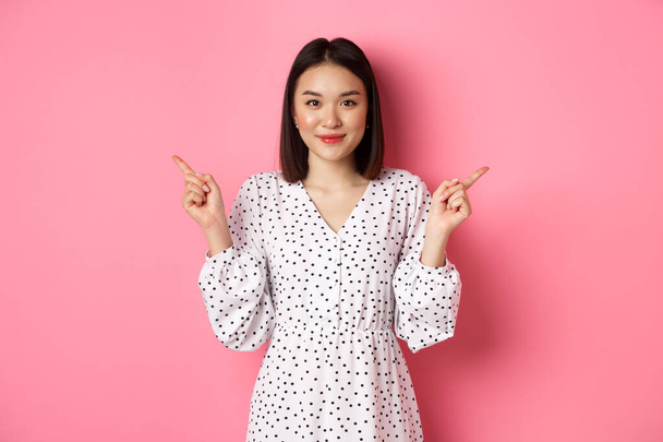 Schöne asiatische Frau trifft die Wahl beim Einkaufen, zeigt mit dem Finger zur Seite und zeigt Varianten, lächelt in die Kamera, steht über rosa Hintergrund - Foto, Bild