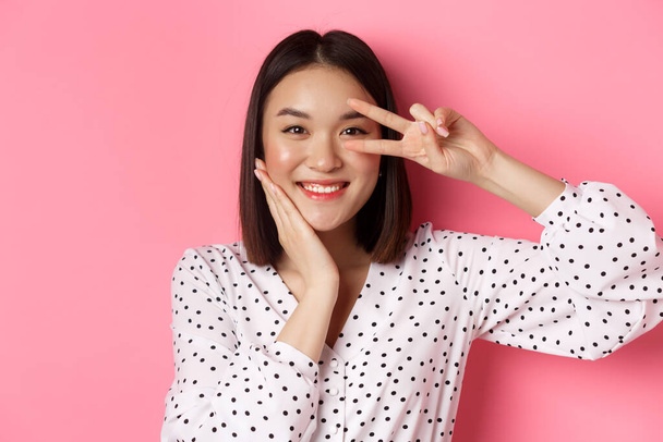 Beauty und Lifestylekonzept. Nahaufnahme einer süßen asiatischen Frau, die Friedenszeichen und rührende Wange zeigt, glücklich in die Kamera lächelt und vor rosa Hintergrund steht - Foto, Bild