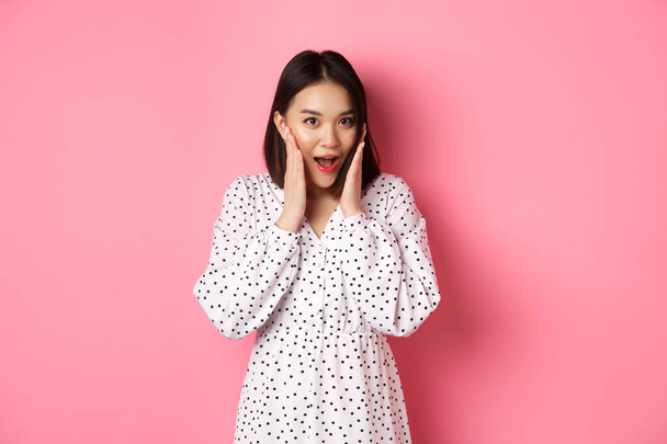 Young asian woman looking intrigued, gossiping and saying wow with curious face, looking interested at camera, standing over pink background - Photo, Image