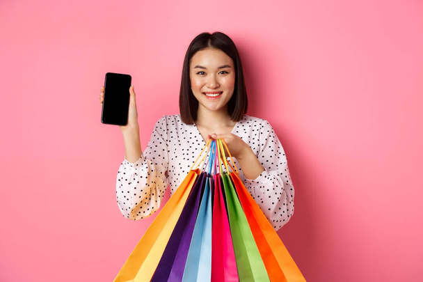 Akıllı telefon uygulaması ve alışveriş poşetleri gösteren çekici Asyalı kadın başvuru yoluyla internetten alışveriş yapıyor, pembe arka planda duruyor. - Fotoğraf, Görsel