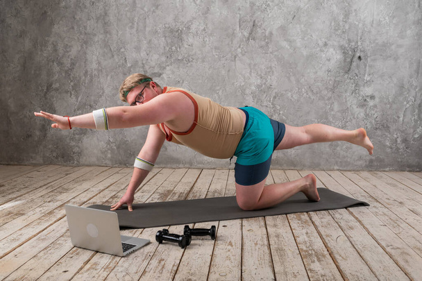 Funny fat man in sportswear doing sports exercises looking into a laptop. home workout  - 写真・画像
