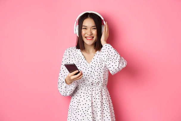 Hermosa mujer asiática escuchando música en auriculares, usando aplicación de transmisión de teléfonos inteligentes y sonriendo, de pie sobre fondo rosa - Foto, Imagen