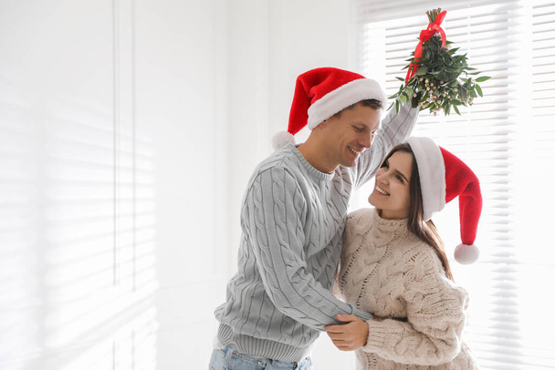 Happy couple in Santa hats under mistletoe bunch at home, space for text - Fotoğraf, Görsel