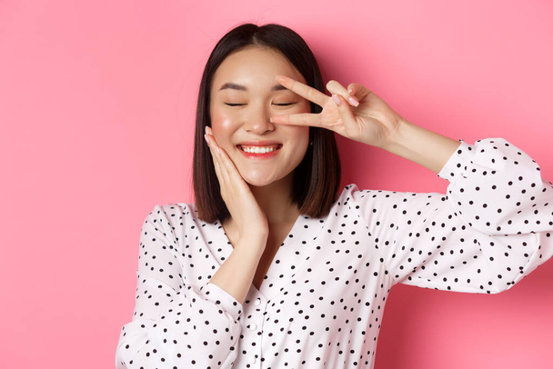Beauty and lifestyle concept. Close-up of pretty asian woman showing peace sign and touching cheek, smiling happy at camera, standing over pink background - Photo, Image