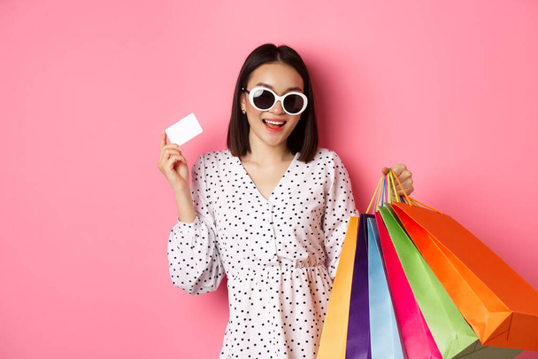 Hermosa mujer asiática en gafas de sol ir de compras, sosteniendo bolsas y mostrando la tarjeta de crédito, de pie sobre fondo rosa - Foto, imagen