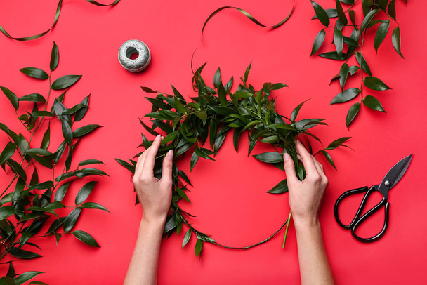 Florist making beautiful mistletoe wreath on red background, top view. Traditional Christmas decor - 写真・画像